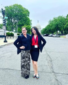 Two women pose on a street wtih the US Capitol visible in the background.
